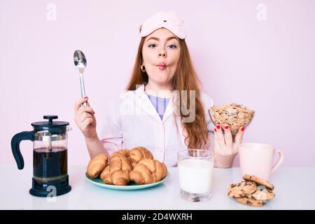 Giovane bella donna rossa seduta sul tavolo facendo colazione a faccia di pesce con bocca e occhi squinting, pazzo e comico. Foto Stock
