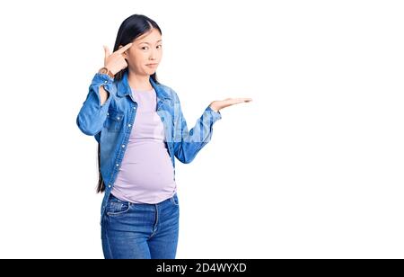 Giovane bella donna cinese incinta aspettandosi bambino confuso e infastidito con palma aperto che mostra lo spazio di copia e che punta il dito alla fronte. Pensi ab Foto Stock