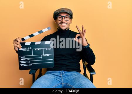 Bell'uomo con tatuaggi reggendo video clapboard seduta sulla sedia regista facendo ok segno con le dita, sorridente amichevole gesturing eccellente symb Foto Stock