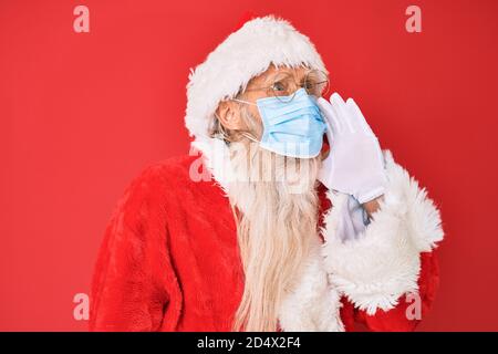 Vecchio uomo anziano che indossa il costume di babbo natale indossando una maschera di sicurezza gridando e gridando forte a fianco con la mano sulla bocca. Concetto di comunicazione. Foto Stock