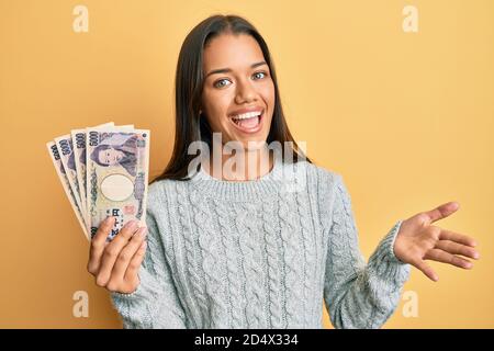 Bella donna ispanica in possesso di 5000 yen giapponesi banconote celebrare il successo con sorriso felice e espressione vincente con mano sollevata Foto Stock