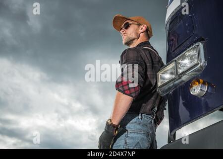 Concetto di settore automobilistico e dei trasporti. Caucasian Trucker driver nei suoi 40 anni davanti al suo retro semi Truck Tractor e Cloudy Sky in Backgro Foto Stock