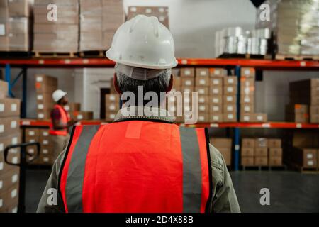 primo piano di lavoratore maschile indossando copricapo e tute arancioni a piedi ai pacchetti in magazzino Foto Stock