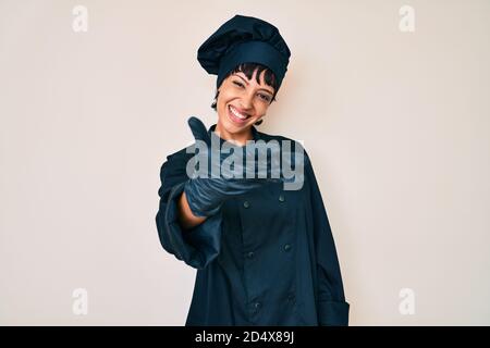 Bella donna brunettte indossare professionista cuoco uniforme sorridente amichevole offrendo handshake come saluto e accogliente. Successo di affari. Foto Stock