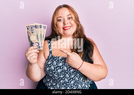 Più taglia giovane donna caucasica che tiene le banconote giapponesi yen sorridenti buon puntamento con mano e dito Foto Stock