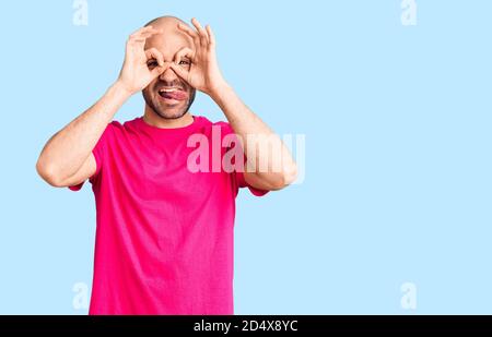 Giovane bell'uomo che indossa una t-shirt casual facendo un gesto ok come binocolo che si stacca la lingua, occhi che guardano attraverso le dita. Espressione pazzesca. Foto Stock