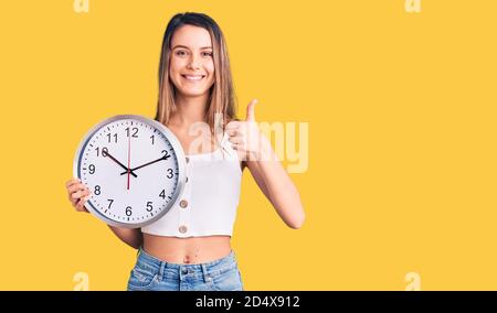 Giovane bella ragazza tenendo grande orologio sorridente felice e positivo, pollice su facendo eccellente e segno di approvazione Foto Stock