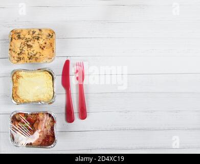 concetto del menu di consegna - tre scatole di alluminio con cibo, forchetta di plastica e coltello isolati su fondo di legno bianco piatto. Take via di organico quotidiano me Foto Stock