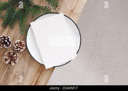 Natale festivo fermo vita. Foglio di carta vuoto, mockup biglietto d'auguri su piastra bianca. Vecchia panca di legno con coni di pino e rames.blurred di abete Foto Stock