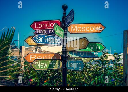 Un colorato cartello che indica le principali città di tutto il mondo, circondato da giovani palme sotto un cielo blu luminoso. Foto Stock