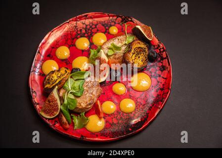 Insalata calda, maiale fritto, fette di fichi, pesche fritte, mirtilli, fette di pomodoro ciliegia, foglie verdi, salsa purea di zucca. Su una targa rossa fatta a mano Foto Stock