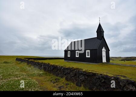 Búðakirkja, una chiesa di legno nero a Budir, Snaefellsnes Peninsular, Islanda. Un muro di pietra circonda l'edificio e le nuvole grigie si riuniscono sopra. Foto Stock