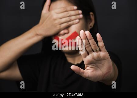 Donna bendata avvolgente bocca con nastro adesivo rosso e mostrare segno di mano smettere di abusare di violenza e abuso su sfondo nero, tratta di esseri umani e ab Foto Stock