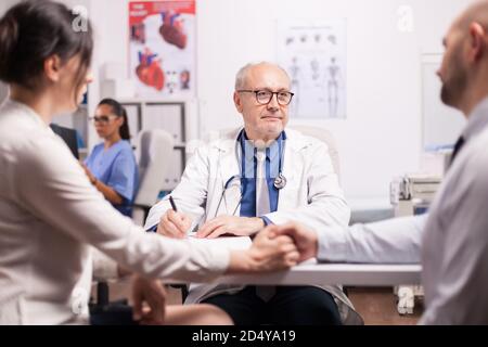 Medico senior che dà cattive notizie a giovani coppie durante il controllo medico in ospedale ufficio. Marito e moglie che tengono le mani. Infermiere che guarda i raggi X del paziente. Foto Stock