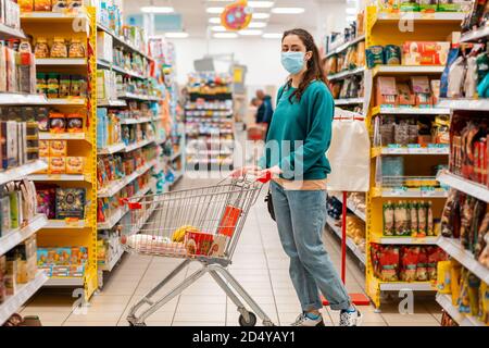 Shopping. Una giovane donna caucasica con una maschera medica sul viso, rotola un carrello della spesa attraverso le corsie del negozio. Vista laterale. Il concetto di buyi Foto Stock