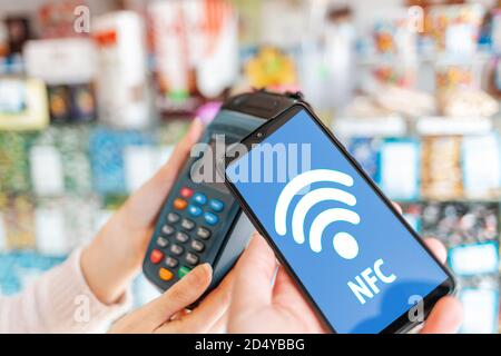 Le mani delle donne sono tenute da un terminale di pagamento e un uomo paga per un acquisto utilizzando uno smartphone.sullo schermo del telefono, la rete wi-fi. Primo piano. La co Foto Stock