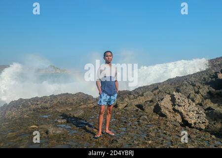 Onde che colpiscono rocce rotonde e spruzzi. Un giovane si erge su una riva rocciosa e le onde si infrangono contro una scogliera Foto Stock