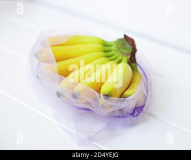 un mazzetto di mini banane in un sacchetto riutilizzabile sopra uno sfondo bianco di legno Foto Stock