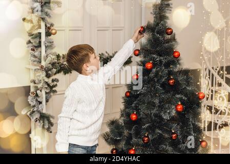 Il piccolo ragazzo carino decora l'albero di Natale con i giocattoli di Capodanno e le palle rosse. Un ragazzo in un pullover bianco lavorato a maglia si siede accanto ai regali e pende palle su un abete artificiale. Concetto di Natale per la famiglia. Foto Stock