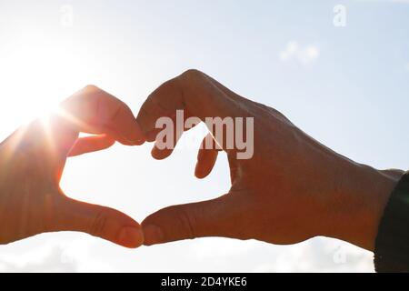 Due mani, maschio e femmina, sono unite a forma di cuore contro il cielo con i raggi del sole all'apertura. Simbolo di una coppia innamorata, Valenti Foto Stock