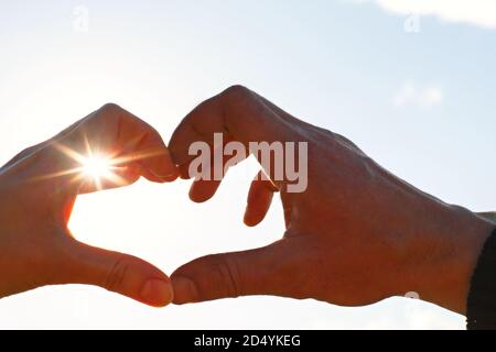 Due mani, maschio e femmina, sono unite a forma di cuore contro il cielo con i raggi del sole all'apertura. Simbolo di una coppia innamorata, Valenti Foto Stock