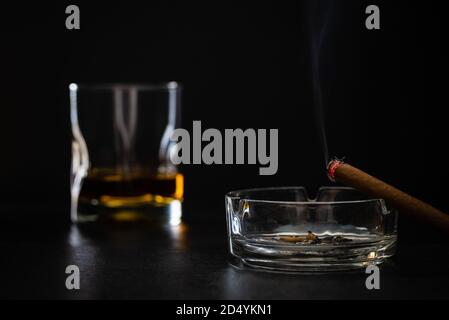 sigaro che brucia con fumo e un bicchiere di whisky sullo sfondo - sfondo nero, fotografia scura Foto Stock