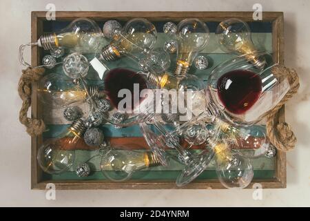 Due bicchieri di vino rosso e retrò ghirlanda di Natale con le lampade accese nel vassoio in legno su marmo bianco dello sfondo. Laici piana, spazio Foto Stock