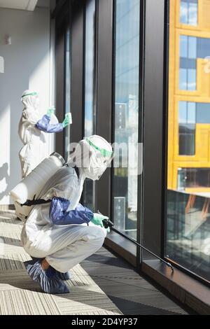 Vista laterale verticale verticale verticale intera lunghezza verticale di due lavoratori che indossano hazmat si adatta alla disinfezione di finestre e pavimenti in un edificio di uffici Foto Stock
