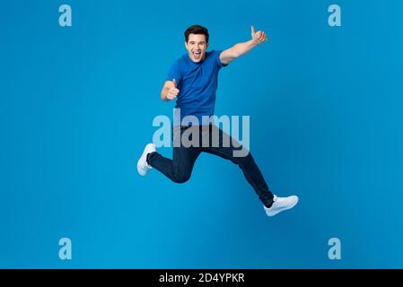 Sorridente bell'uomo americano saltando gioiosamente e facendo doppio pollice gesto verso l'alto isolato su sfondo blu dello studio Foto Stock