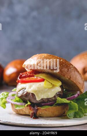 Close up in casa Hamburger classico nel frumento bun con carne di manzo e costolette di vitello, il formaggio fuso e le verdure su ceramica bianca sulla scheda blu grigio nella tabella. Foto Stock