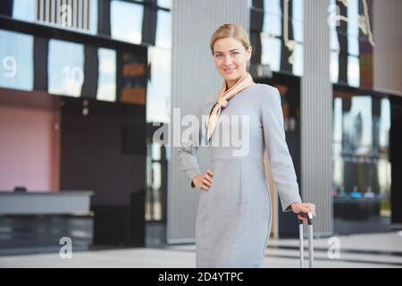 Ritratto di elegante donna bionda con valigia guardando la macchina fotografica e sorridendo mentre si posa in aeroporto, copia spazio Foto Stock
