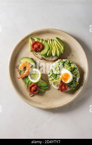 Varietà di panini vegetariani con fette di avocado, pomodori secchi, uovo, gamberetti, rucola servita su piastra in ceramica oltre bianco grigio maculato backgr Foto Stock