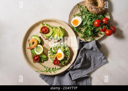 Varietà di panini vegetariani con fette di avocado, pomodori secchi, uovo, gamberetti, rucola servita su piastra in ceramica oltre bianco grigio maculato backgr Foto Stock