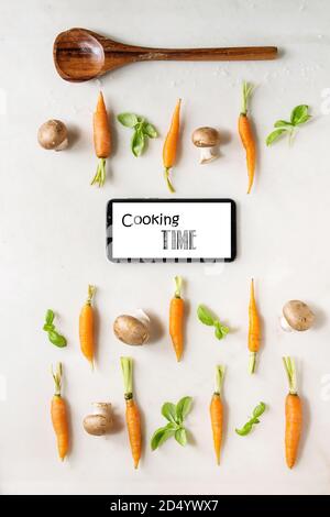 Il basilico fresco erbe con giovani mini carote e funghi champignon in righe, il cucchiaio di legno e lo smartphone su marmo bianco dello sfondo. Appartamento laico, spazio. Foto Stock
