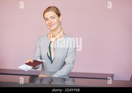 Ritratto in vita di un elegante assistente di volo dietro il banco del check-in e sorridente presso la fotocamera che consegna i biglietti al passeggero, copy space Foto Stock