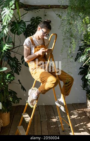 Donna giardiniere in tute arancioni seduta sulla scala nella sua casa verde, riposando, utilizzando smartphone, leggendo messaggio al telefono. Pausa durante il lavoro Foto Stock