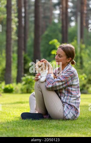 Donna felice in camicia controllata abbracciando e abbracciando con tenerezza e amore domestico zenzero gatto, seduta su erba all'aperto in giornata di sole. Amore per la a Foto Stock
