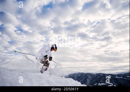 Vista laterale dello sciatore maschile che corre lungo pendii innevati su sci sotto il bel cielo nuvoloso. Uomo freerider sciare su neve fresca in alta montagna d'inverno. Concetto di sci e di svago attivo. Foto Stock