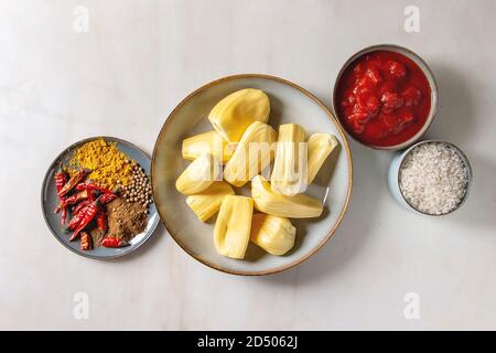 Gli ingredienti per cucinare vegan curry. Raw maturo pelato jackfruit con il bianco riso crudo, tagliate a pezzetti i pomodori e spezie in ceramica bocce su bianco marbl Foto Stock