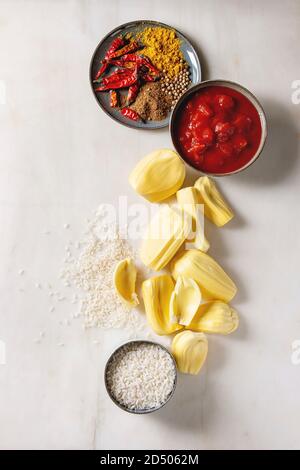 Gli ingredienti per cucinare vegan curry. Raw maturo pelato jackfruit con il bianco riso crudo, tagliate a pezzetti i pomodori e spezie in ceramica bocce su bianco marbl Foto Stock