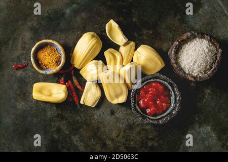 Gli ingredienti per cucinare vegan curry. Raw maturo pelato jackfruit con il bianco riso crudo, tagliate a pezzetti i pomodori e spezie in ciotole di ceramica su metallo scuro Foto Stock