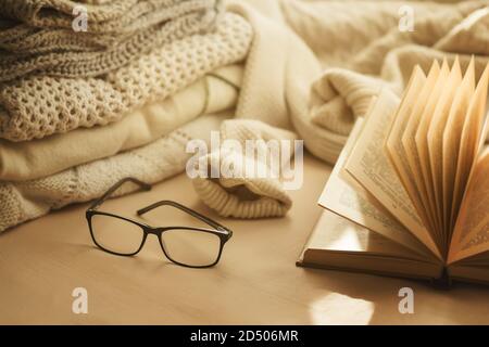 I pullover morbidi e caldi sono accatastati sul letto, insieme a un libro aperto e occhiali neri al sole. Comfort a casa. Processo di lettura. Foto Stock