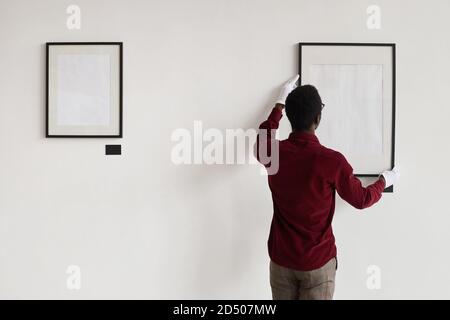 Vista posteriore di un uomo afro-americano appeso cornici vuote sulla parete mentre si pianifica galleria d'arte o mostra, copia spazio Foto Stock