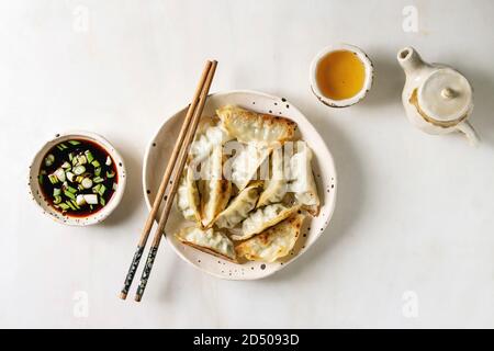 Fritti gnocchi asiatici Gyozas potstickers in bianco piastra ceramica servita con bacchette, ciotola di soia Salsa di cipolla, teiera su marmo bianco dello sfondo. Foto Stock