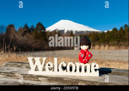 Bella bambola tradizionale giapponese kokeshi vestito in rosso kimono sopra la parola 'Benvenuto'. Sfondo con Monte Fuji. Shot orizzontale. Foto Stock