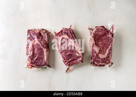 Tre materie non cotte le bistecche di manzo in confezionato sotto vuoto in fila su marmo bianco dello sfondo. Pronto per il sous vide la cottura. Laici piana, spazio Foto Stock