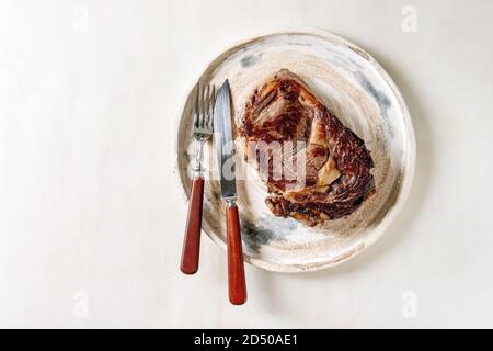 Grigliate di arrosto di mezzo raro bistecca di manzo servita nella piastra in ceramica con posate su marmo bianco dello sfondo. Laici piana, spazio Foto Stock