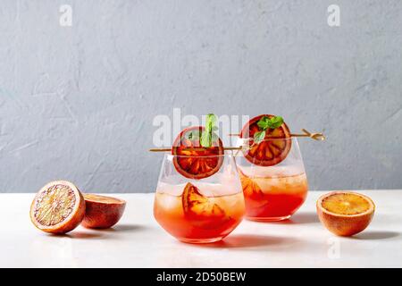 Arancio sanguigno cocktail ghiacciato in bicchieri, decorata da fetta di arance e menta fresca su spiedini, servita su marmo bianco tavola con muro grigio in backgr Foto Stock