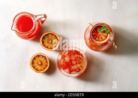 Arancio sanguigno cocktail ghiacciato in bicchieri, decorata da fetta di arance e menta fresca su spiedini, servito con una brocca di succo in marmo bianco sullo sfondo Foto Stock