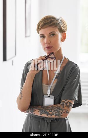 Verticale vita in su ritratto di donna tatuata elegante guardando macchina fotografica mentre ci si trova in galleria d'arte o in museo Foto Stock